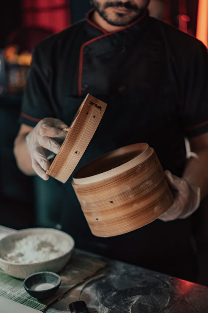 pexels-photo-8092561 Chef in black uniform preparing food with a bamboo steamer in a dimly lit kitchen.